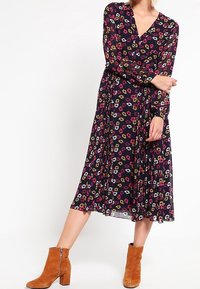 Das florale Wickelkleid hat einen marineblauen Hintergrund mit mehrfarbigen Blumen, langen Ärmeln und einem plissierten Rock. Kombiniert mit braunen Ankle Boots.