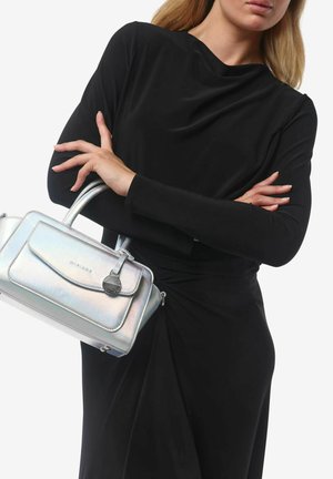 Borsa a tracolla argentata metallic, con una texture liscia, forma strutturata, pattina frontale e un charm decorativo. Indossata con un abito nero a maniche lunghe.