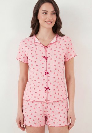 Mujer con pijama rosa de manga corta con pequeños estampados florales rojos y lazos rojos decorativos en la parte delantera, de pie frente a un fondo blanco.