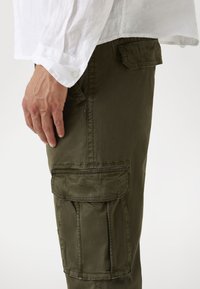 Olijfgroene cargo broek van stevig materiaal, met meerdere zakken, zichtbare stiksels en een relaxte pasvorm. Bovenop is een witte linnen shirt gedragen.