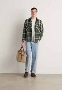 Camicia di flanella a quadri verde e nera, t-shirt bianca, jeans azzurri chiari, scarpe marroni, reggendo una borsa beige con dettagli marroni.
