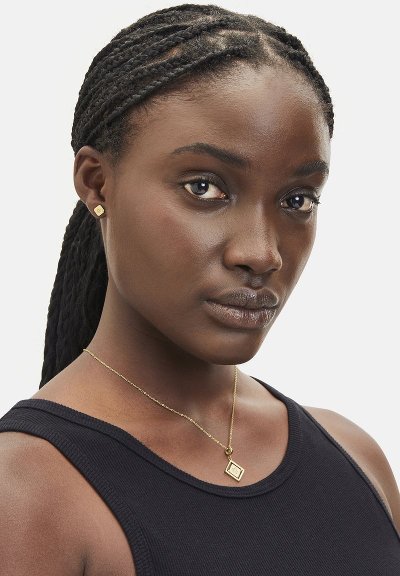 Gold pendant necklace with a square charm and geometric design, worn over a black ribbed tank top, paired with small gold square stud earrings.