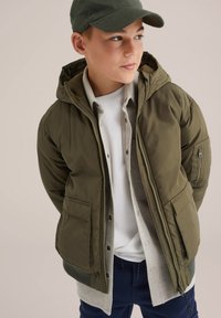 Olivno zelena puhasta jakna s kapuco, dvema velikima sprednjima žepoma in z zadrgo, nosena čez svetlo sivo majico in belo majico s kratkimi rokavi.