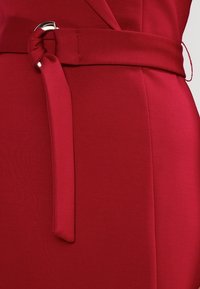 Czerwony materiał sukienki o gładkiej fakturze, z węzłem w talii i detalami ze srebrnego metalu. Zbliżenie podkreśla fason i kształt.