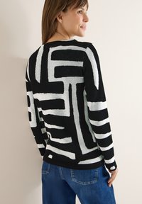 Czarny i biały sweter dzianinowy w wzory z abstrakcyjnymi liniami. Posiada długie rękawy i okrągły dekolt, zestawiony z niebieskimi dżinsami.