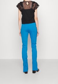 Haut noir avec des manches en voile à volants, dos en forme de clé, associé à un pantalon ajusté bleu vif. Texture lisse, design taillé et accents minimalistes.