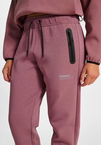 Lila sportiga joggers med en ficka med dragkedja, dragsko i midjan och en slät textur. Försedd med "hummel"-logotypen på nedre benet.