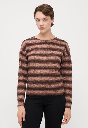 Vrouw in een bruin en donkerbruin gestreept lange mouw sweater en zwarte broek, staande tegen een effen lichte achtergrond.
