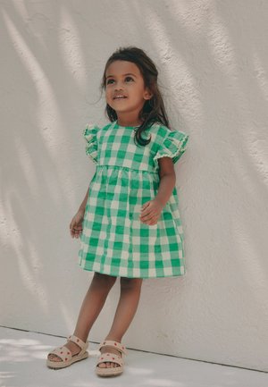 Una giovane ragazza sta in piedi contro un muro bianco strutturato, indossa un vestito a quadri verde e bianco e sandali beige con motivi di fragole.