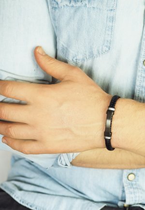Black braided bracelet with a flat metal clasp, worn on a light-skinned wrist, against a light blue denim shirt.