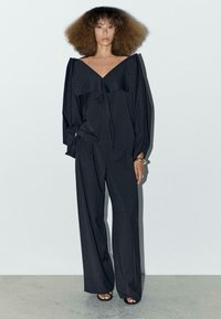 Czarna, oversize bluzka z dekoltem w serek i detalami w postaci falbanek, w zestawieniu z szerokimi spodniami. Gładka tekstura; minimalistyczny design.