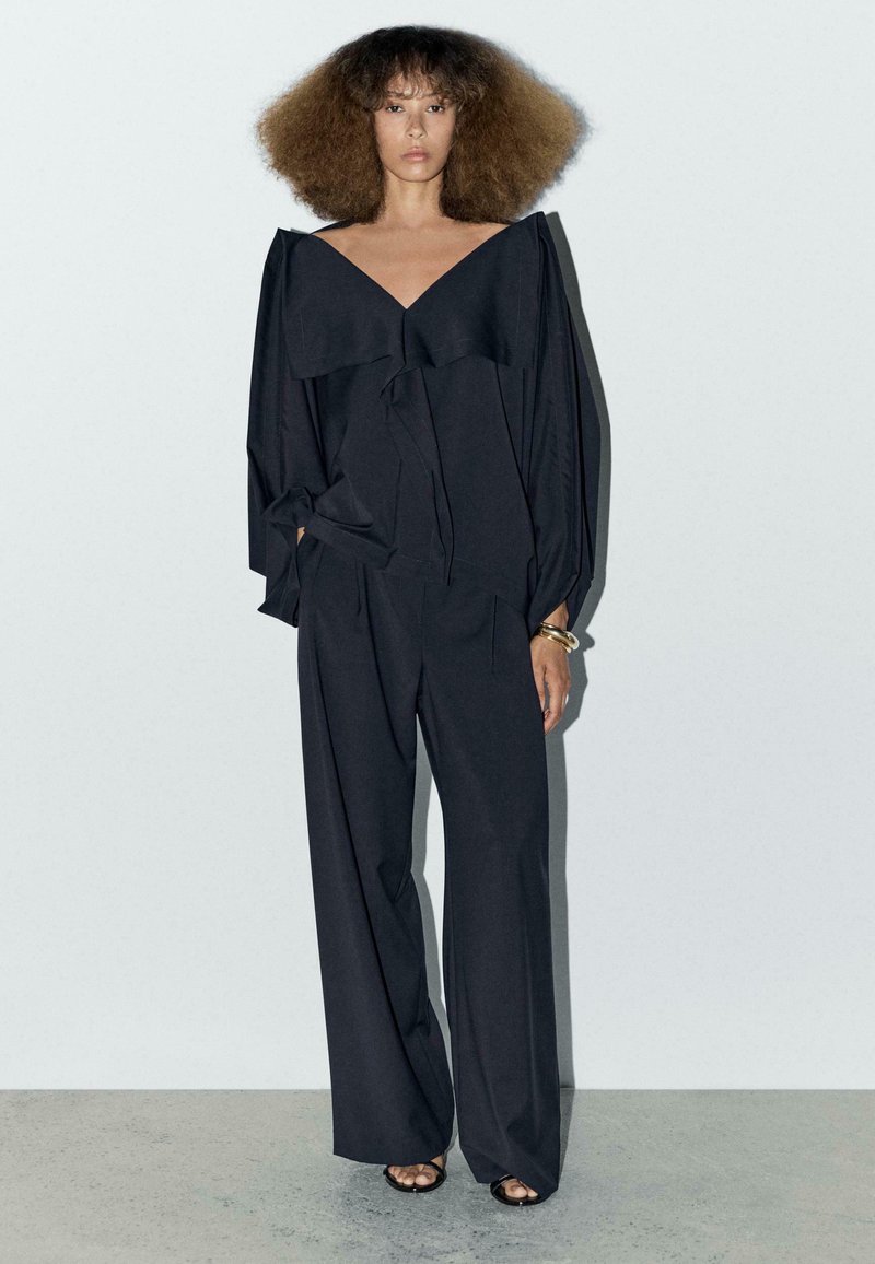 Czarna, oversize bluzka z dekoltem w serek i detalami w postaci falbanek, w zestawieniu z szerokimi spodniami. Gładka tekstura; minimalistyczny design.