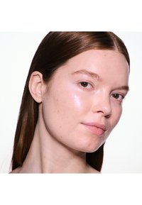 Gros plan d'un visage avec une peau lisse et claire ; sous-tons rose pâle et éclat naturel. Cheveux bruns longs et raides ; arrière-plan neutre.