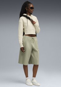 Geknipte beige hoodie met koord, gecombineerd met lichtgroene shorts en witte sneakers. Zwarte zonnebril en textuurhaar accentueren de look.