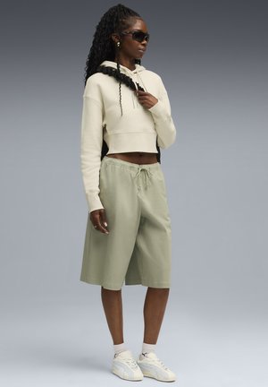 Geknipte beige hoodie met koord, gecombineerd met lichtgroene shorts en witte sneakers. Zwarte zonnebril en textuurhaar accentueren de look.