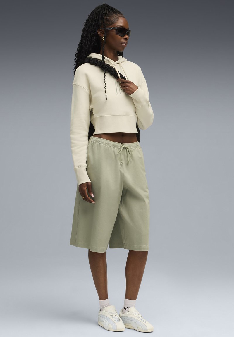 Geknipte beige hoodie met koord, gecombineerd met lichtgroene shorts en witte sneakers. Zwarte zonnebril en textuurhaar accentueren de look.