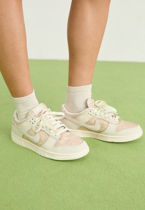 Piernas con calcetines blancos hasta el tobillo y zapatillas Nike beige claro con suelas blancas sobre una alfombra verde.