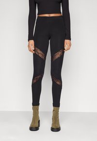 Zwarte leggings met kanten accenten in een zigzagpatroon op de dijen, gecombineerd met olijfgroene enkellaarsjes. Glad stoffen afwerking.