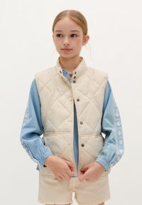 Gilet matelassé crème avec col montant, fermetures à pression, porté sur une blouse bleu clair avec des manches brodées, associé à un short beige.