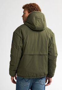 Grüne gefütterte Jacke mit Kapuze, im lässigen Schnitt, sichtbaren Nähten und einem Rückenschlappdetail. Getragen mit blauen Jeans.