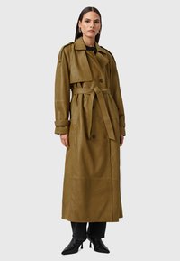 Olijfgroene leren trenchcoat met dubbele knopen, een ceintuur in de taille, brede kraag en lange mouwen. Gecombineerd met donkere jeans en zwarte hakken.