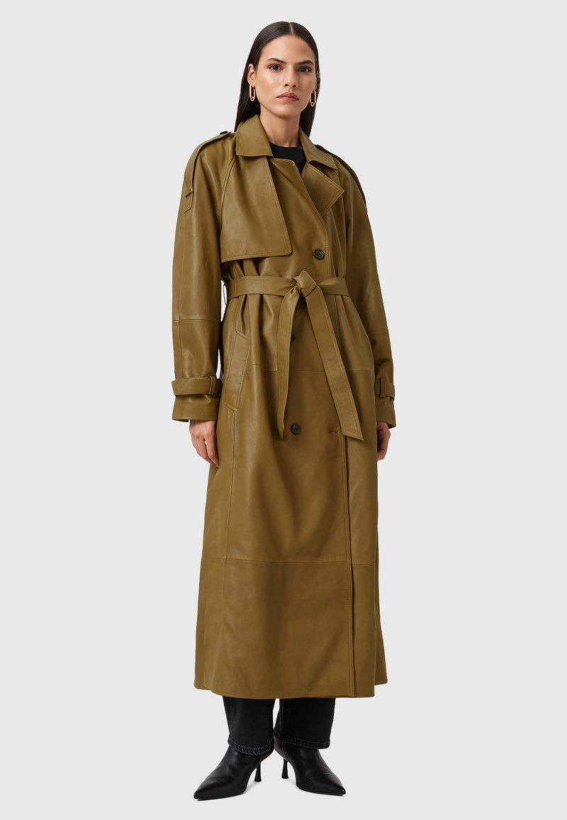 Olijfgroene leren trenchcoat met dubbele knopen, een ceintuur in de taille, brede kraag en lange mouwen. Gecombineerd met donkere jeans en zwarte hakken.