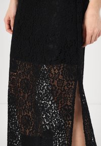 Rochie neagră din dantelă cu modele florale, o fenta înaltă și secțiuni transparente, oferind un design texturat și complex.