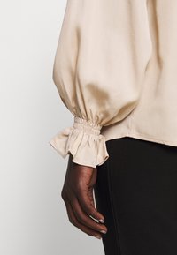 Blouse beige à manches longues bouffantes, poignets froncés et texture lisse. Présente une coupe ample et une légère brillance.