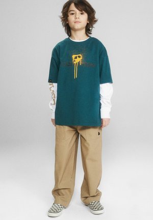 Chłopiec o średniej długości włosach nosi turkusowy T-shirt z nadrukiem na białej koszulce z długim rękawem, beżowe spodnie i w kratkę buty wsuwane, stoi prosto.