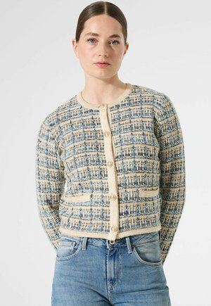 Femme portant un cardigan texturé bleu et beige à boutons avec des poches avant, associé à un jean taille haute bleu clair sur fond uni.