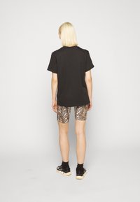 Vaalea henkilö, joka katsoo pois, pukeutuneena löysään mustaan t-paitaan, käärmeennahkakuosisiiin pyöräilyshortseihin, mustiin sukkiin ja musta-valkoisiin lenkkareihin tasaisella taustalla.