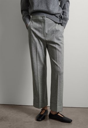 Persoon draagt grijze, op maat gemaakte broek met omslagen en een grijze trui, handen in de zakken en zwarte platte schoenen met enkelbandjes.