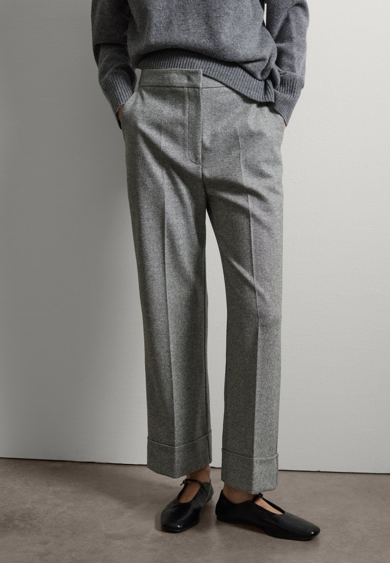 Personne portant un pantalon gris sur mesure avec revers et un pull gris, mains dans les poches, et des chaussures plates noires avec des brides à la cheville.