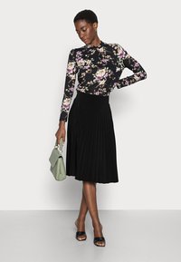 Jupe plissée noire associée à un haut à manches longues à fleurs avec un fond noir et des nuances de rose et de vert, complétée par un sac vert clair.