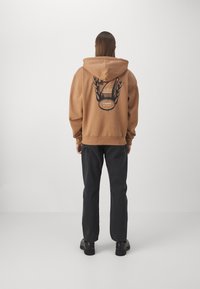 Sudadera marrón con un diseño gráfico en la parte trasera que presenta llamas y el texto "7 DAYS ACTIVE". Combinada con jeans oscuros y zapatos negros.