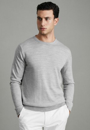 Pulover lung cu mâneci lungi de culoare gri deschis, cu guler rotund, un material texturat și mansete și tiv cu nervuri, asortat cu pantaloni albi.