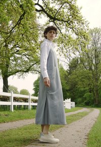 Abito grigio senza maniche con una fessura laterale, caratterizzato da un colletto bianco e una tasca. La modella indossa scarpe da ginnastica chiare. Sfondo verde naturale.