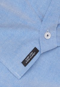 Lichtblauw, gestructureerde stof kraag van een shirt met een witte knop. Een zwart label met de tekst "IEDERE STAP EEN NIEUWE REIS" staat op de kraag.