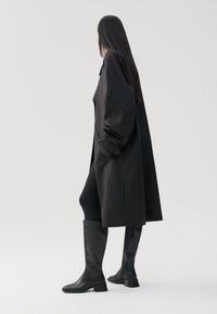 Abrigo negro de corte amplio, con puños abotonados y bolsillos laterales, combinado con botas de cuero negro hasta la rodilla. Textura suave.