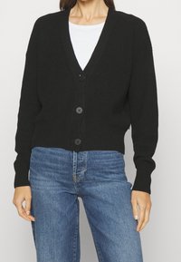 Femme portant un cardigan noir à boutons sur un top blanc, associé à un pantalon en jeans bleu taille haute.