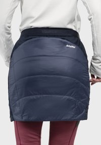 Marineblauer isolierter Rock mit steppoptik, ausgestattet mit einem schwarzen elastischen Bund und einem Logo-Akzent auf der Rückseite. Kombiniert mit burgunderfarbenen Hosen.