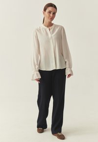 Blouse beige clair à manches longues évasées avec des boutons, associée à un pantalon large bleu marine foncé et des baskets marron.