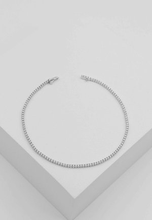 Silbernes Tennisarmband mit kleinen runden klaren Steinen in einer einzelnen Reihe, liegend auf einer weißen geometrischen Oberfläche.