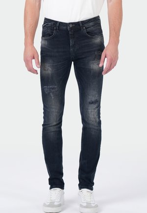 Vorderansicht einer Person, die ausgewaschene schwarze Slim-Fit-Jeans mit abgenutzten Stellen, ein weißes Hemd und weiße Turnschuhe vor einem einfarbigen Hintergrund trägt.