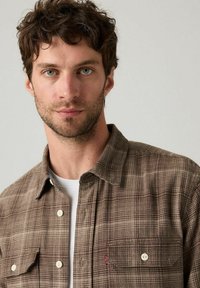 Giovane uomo con capelli ricci castani che indossa una camicia a quadri marrone sopra una t-shirt bianca, guarda direttamente verso la fotocamera.