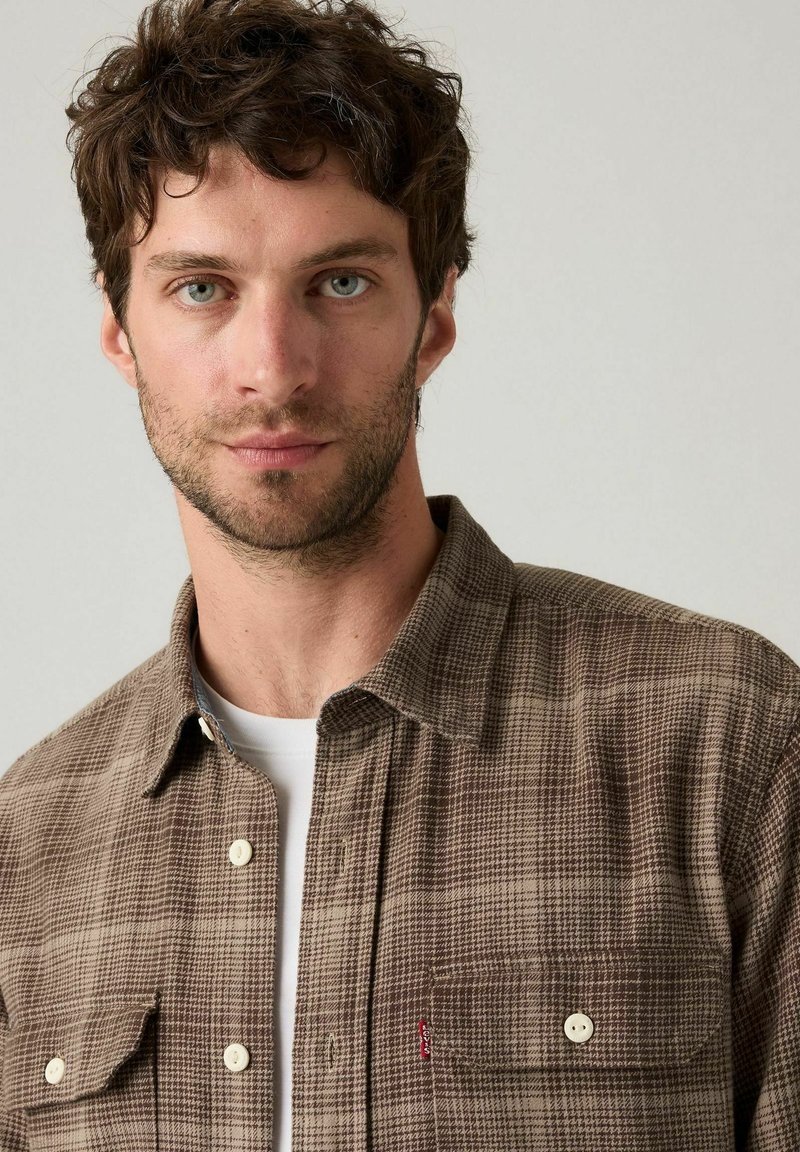 Junger Mann mit lockigem braunem Haar, der ein braunes kariertes Hemd über einem weißen T-Shirt trägt und direkt in die Kamera schaut.