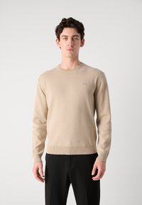 Beige kötött pulóver kerek nyakkivágással, bordás mandzsettákkal és szegéllyel, mellkasán egy kis logóval. Sima textúrájú és testhezálló fazon.
