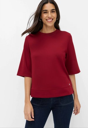 Smilende kvinde med langt mørkt hår iført gyldne hoop-øreringe, en rød top med korte ærmer og mørkeblå jeans, stående mod en hvid baggrund.