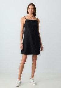 Femme debout dans une robe noire sans manches avec de fines bretelles et des baskets blanches devant un fond gris clair uni.