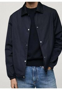 Veste légère bleu marine avec un col classique, fermeture à boutons-pression, poignets élastiques et texture de tissu lisse. Portée sur un pull noir.